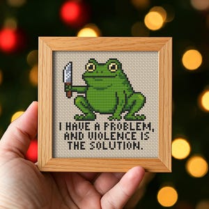 Könnte beinhalten: Kreuzstich-Kunstwerk in einem Holzrahmen. Es zeigt einen grünen Frosch, der ein Messer hält, mit dem Text: „ICH HABE EIN PROBLEM, UND GEWALT IST DIE LÖSUNG.“ Der Hintergrund ist ein verschwommener Weihnachtslicht-Anzeiger.