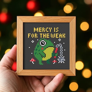 May include: Cross-stitch artwork in a wooden frame. The design features a green frog with a yellow belly holding a chain, with the text "MERCY IS FOR THE WEAK" in yellow. The background is dark gray.