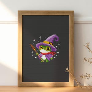 May include: Cross-stitch art of a green frog wearing a purple witch hat and scarf, holding a broom. The frog is set against a dark gray background, framed in a light brown wooden frame. The artwork has a whimsical, Halloween theme.