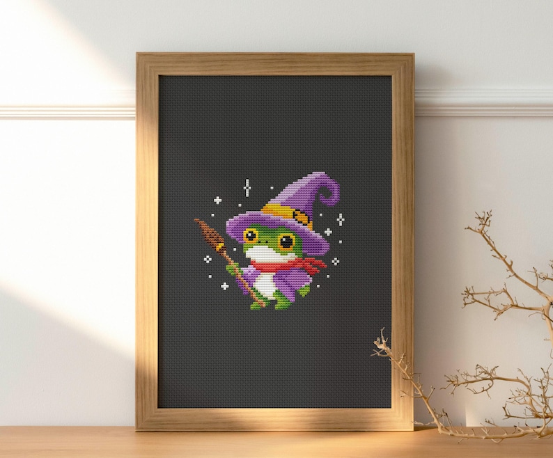 May include: Cross-stitch art of a green frog wearing a purple witch hat and scarf, holding a broom. The frog is set against a dark gray background, framed in a light brown wooden frame. The artwork has a whimsical, Halloween theme.