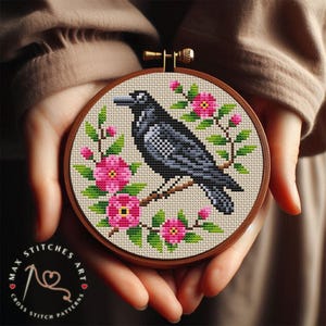 Puede incluir: Un patrón de punto de cruz de un cuervo negro con flores rosas sobre un fondo blanco. El patrón está enmarcado en un aro de madera.