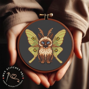 May include: Cross-stitch art featuring a cat with butterfly wings, in a wooden embroidery hoop. The cat is brown and tan with blue eyes, and the wings are green and yellow. The background is dark gray.