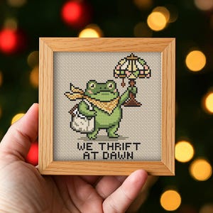 May include: A square wooden frame containing a cross-stitch embroidery of a green frog wearing a yellow scarf and carrying a white bag. The frog holds a stained-glass style lamp. The text WE THRIFT AT DAWN is stitched in black below the frog.