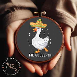 May include: A cross-stitch embroidery hoop featuring a white goose wearing a sombrero, with the text "ME GOOSE-TA" below. The goose is walking with its feet in an orange color. The background is dark gray, and the hoop is held in a person's hands.