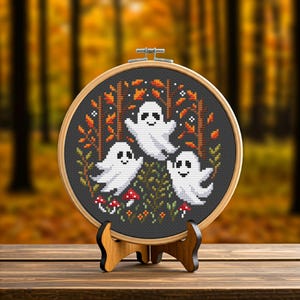 May include: A cross-stitch embroidery hoop featuring three smiling white ghosts in a forest scene with orange and red leaves, trees, and mushrooms. The embroidery is set in a wooden hoop and displayed on a wooden stand.