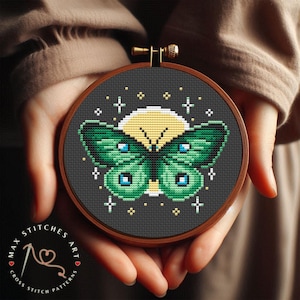 Könnte beinhalten: Ein Kreuzstichmuster mit einem grünen Schmetterling mit gelben Akzenten, umgeben von einem gelben Mond und weißen Sternen auf einem dunkelgrauen Hintergrund.