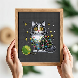 Op de afbeelding: Kruissteekpatroon van een zwart-witte kat die kerstverlichting draagt en een groene kerstbal vasthoudt.