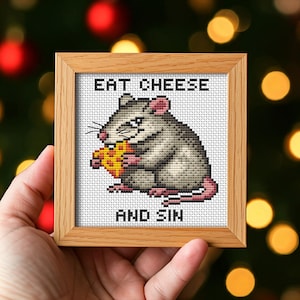 May include: A framed cross-stitch featuring a gray rat eating a piece of yellow cheese. The text "EAT CHEESE" is above the rat, and "AND SIN" is below. The frame is made of light brown wood.