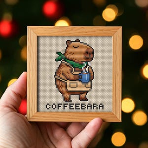 May include: A framed cross-stitch depicting a brown capybara wearing a green scarf and apron, holding a blue mug. The text "COFFEEBARA" is stitched below the capybara. The frame is made of light brown wood.