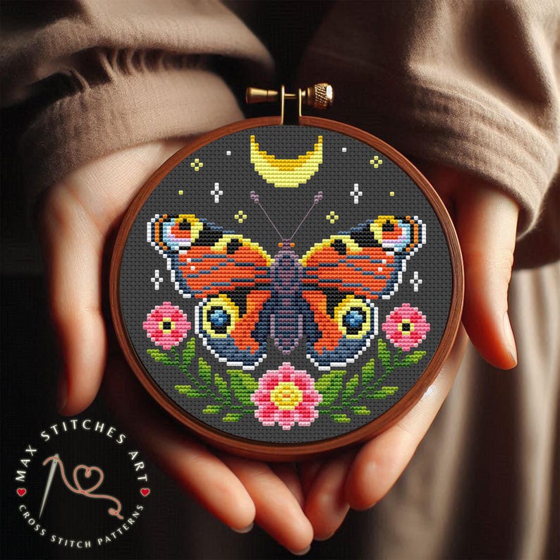 May include: A cross stitch pattern featuring a colorful butterfly with a crescent moon and stars on a dark background. The butterfly has orange, black, and white wings with a pink and yellow flower on each side.