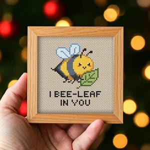 May include: A square light wood frame containing a cross-stitch embroidery of a yellow and black bee holding a green leaf. The text I BEE-LEAF IN YOU is stitched in black below the bee on a cream-colored fabric background within the wooden frame.