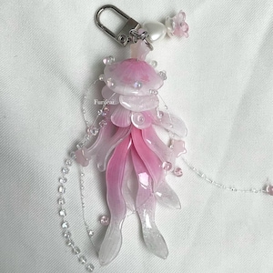 Puede incluir: Llavero decorativo con forma de medusa, con pétalos translúcidos rosas y blancos. Tiene un cierre plateado y está adornado con cuentas transparentes y una pequeña flor. La palabra "Furifeai" está impresa en el lateral.