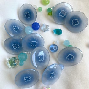 Può includere: Una collezione di 10 cabochon in vetro blu chiaro con un motivo a fiore bianco. I cabochon sono disposti a semicerchio su una superficie bianca. Ci sono anche diverse piccole perline di vetro blu, verdi e iridescenti sparse intorno ai cabochon.