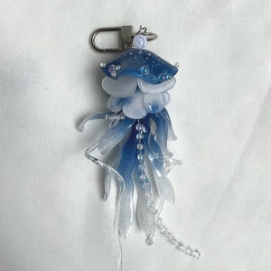May include: A blue and white jellyfish keychain with a silver clasp. The jellyfish has a domed head with white dots and iridescent accents. The body is layered with translucent petals and dangling beaded strands.