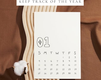 Könnte beinhalten: Eine weiße Kalenderseite für Januar mit dem Text "KEEP TRACK OF THE YEAR". Der Kalender hat ein minimalistisches Design mit der Nummer "01" und einer Sternengrafik. Der Kalender befindet sich auf einem gewellten Holzgrund.