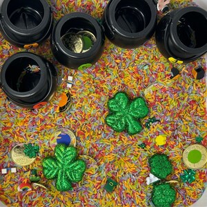 May include: A white plastic container filled with colorful rice and small toys, including shamrocks, coins, and black pots.