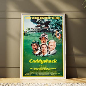 May include: A framed Caddyshack movie poster featuring a large gopher illustration above a golf course scene with portraits of Bill Murray, Chevy Chase, and Ted Knight. The text reads Some People Just Don't Belong and Caddyshack with cast credits.