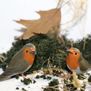 May include: Two small wooden robin figurines with orange breasts and gray bodies are perched on a bed of green moss. The robins are facing forward with their heads tilted slightly to the side.