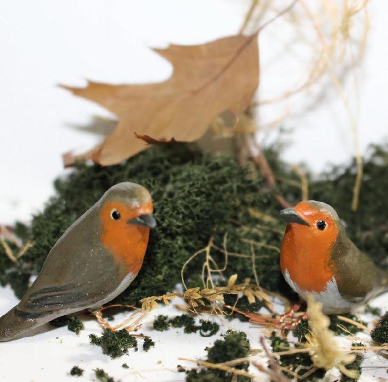 May include: Two small wooden robin figurines with orange breasts and gray bodies are perched on a bed of green moss. The robins are facing forward with their heads tilted slightly to the side.
