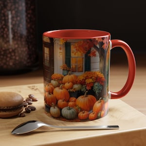 May include: A ceramic mug with a red interior and handle, featuring a fall-themed illustration. The design depicts a house with pumpkins, flowers, and autumn foliage. The mug is on a wooden surface with a spoon and a macaron.