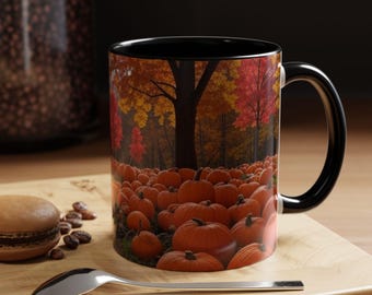Op de afbeelding: Koffiemok met zwarte handgreep met een herfsttafereel van pompoenen en bomen. De mok toont een veld met oranje pompoenen onder een boom met kleurrijk herfstblad. Een lepel en een gebakje staan op de voorgrond.