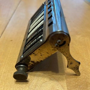 May include: Vintage metal desk calculator with a silver finish. The calculator has multiple number dials with black numbers on a white background. The calculator is angled and has a small, adjustable foot for support. The calculator is on a wooden surface.