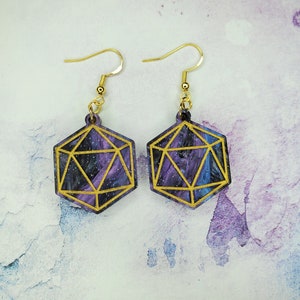 May include: A pair of gold dangle earrings with a galaxy-themed design. The earrings are shaped like a 20-sided die, with a gold outline and a purple, blue, and black galaxy design inside.