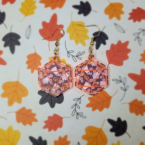 May include: A pair of dangle earrings featuring a gold-colored hook and a translucent orange and white die with a ghost and pumpkin pattern.