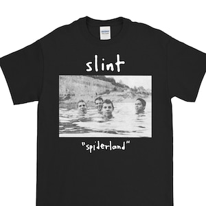 Puede incluir: Camiseta negra con el nombre de la banda "slint" en blanco sobre una imagen en blanco y negro de cuatro personas en el agua. La palabra "spiderland" está escrita debajo en blanco.