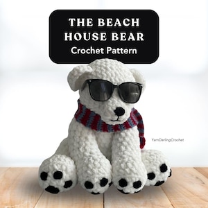 Op de afbeelding: Een witte gehaakte beer met een zwarte zonnebril en een rood, wit en blauw sjaaltje. De beer heeft zwarte poten en een zwarte neus. De tekst "THE BEACH HOUSE BEAR Crochet Pattern" staat bovenaan.