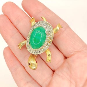 May include: A gold-toned turtle brooch with a large oval green stone center, surrounded by clear crystals. The turtle's head has a small red stone eye. The brooch is a vintage jewelry piece.