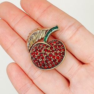 May include: A gold-tone cherry brooch with red rhinestone accents. The stem of the cherry is green enamel.