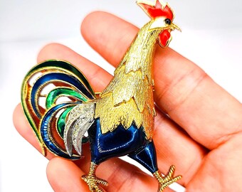 May include: A gold-toned rooster brooch with red, blue, green, and white enamel detailing. The rooster has a textured gold body, a red comb, and a blue base. The brooch is held in a hand.