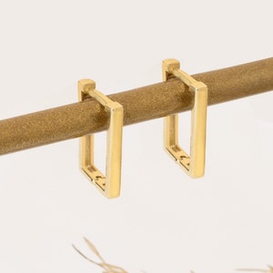 May include: A pair of gold square hoop earrings hanging from a wooden dowel rod. The earrings are simple and minimalist in design.