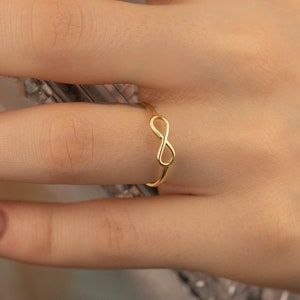 May include: A gold infinity ring on a finger with nude nail polish.