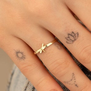 May include: A gold ring with a dragonfly design. The ring is worn on the index finger of a hand with several small tattoos.