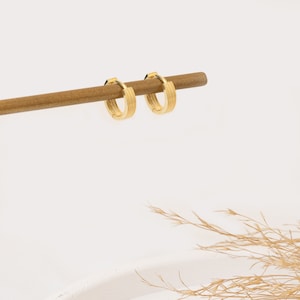 Könnte beinhalten: Zwei goldfarbene Creolen mit einem strukturierten, geriffelten Design. Die Ohrringe werden auf einem dünnen, braunen Holzstab vor einem weißen Hintergrund präsentiert. Ein klassisches Schmuckstück.