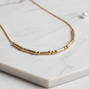 May include: A delicate gold necklace with a series of small, round beads and cylindrical bars. The necklace is displayed on a white marble surface, with a single pearl visible in the background. The jewelry has a simple, elegant design.
