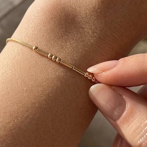 May include: A delicate gold-toned chain bracelet featuring a series of small cylindrical and spherical beads arranged in a pattern that resembles Morse code. The bracelet is thin and rests against the skin, showcasing a metallic finish.