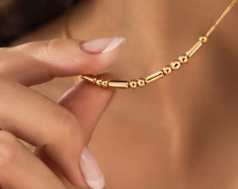 以下が含まれることがあります： ゴールドトーンのネックレス。繊細なチェーンの中央に、磨かれた小さな球状のビーズと細長い円筒形のバーが交互に並び、繰り返しのパターンを形成しているデザイン。