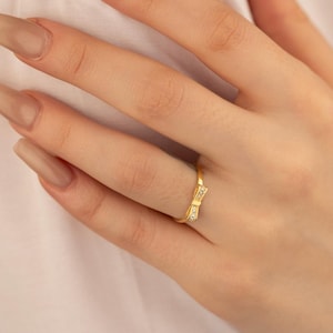 May include: A gold-colored ring featuring a bow design with small, clear gemstones. The ring is on a finger, showcasing its delicate details and elegant style. The bow is the focal point, adding a touch of charm to the jewelry.