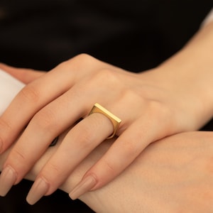 May include: A gold-colored ring with a rectangular design. The ring is worn on a finger, showcasing its geometric shape and polished finish. The ring's design is simple yet modern, making it a versatile accessory.