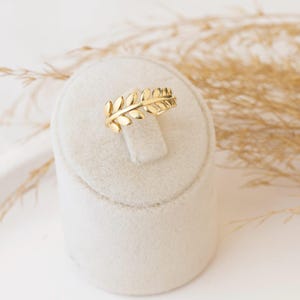 May include: A gold-colored ring featuring a leaf design. The ring is displayed on a white, velvet-covered stand. The leaves are detailed and arranged in a band around the ring. The background includes dried, tan-colored plants.