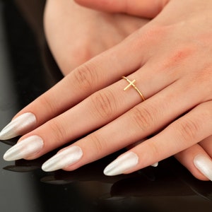 May include: A delicate gold-colored ring featuring a simple cross design. The ring is thin and minimalist, with the cross positioned on the top of the finger. The ring is shown on a hand with long, pearlescent white nails.