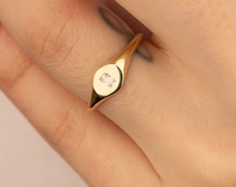 May include: A gold-colored ring with a rectangular, clear gemstone set in a circular bezel. The ring has a smooth, polished finish and a slender band. The gemstone is approximately 0.1 inches in length.