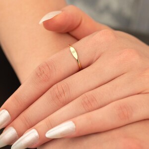 May include: A gold-colored ring with an oval-shaped face, featuring the letter "B" engraved on it. The ring is delicate and thin, with a polished finish. The ring is worn on a finger.