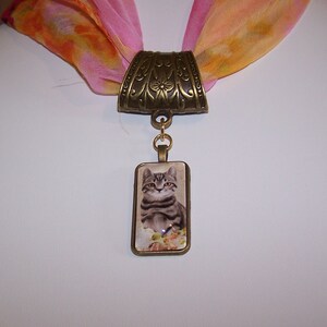 May include: A bronze scarf ring with an ornate design and a rectangular pendant featuring a gray tabby cat with flowers.