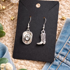 May include: Silver-toned cowboy-themed earrings. One earring features a cowboy hat design with a central clear stone, while the other showcases a cowboy boot. The earrings hang from silver hooks and are displayed on a black card.