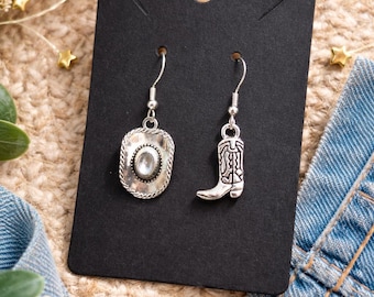May include: Silver-toned cowboy-themed earrings. One earring features a cowboy hat design with a central clear stone, while the other showcases a cowboy boot. The earrings hang from silver hooks and are displayed on a black card.
