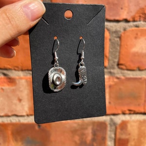 May include: Silver-toned cowboy-themed earrings. One earring is a cowboy hat, the other a cowboy boot. The earrings are displayed on a black card against a brick background.